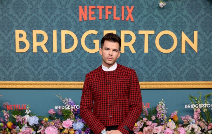 Luke Newton at a Netflix Bridgerton event, wearing a stylish plaid jacket and black pants, standing in front of a floral arrangement and promotional backdrop