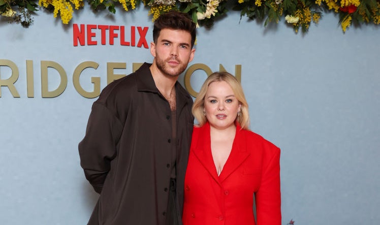 Nicola Coughlan in a red blazer-dress and red heels, and Luke Newton in a dark suit, posing at the Netflix Bridgerton event with a floral backdrop