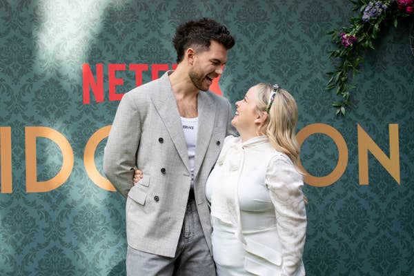 Luke Newton and Nicola Coughlan smiling at each other at a Bridgerton event, Luke in a blazer and Nicola in a white outfit