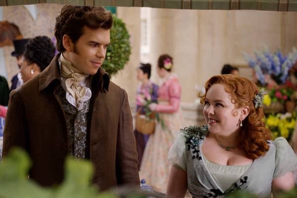 Nicola Coughlan and Luke Newton in period costumes on the set of a historical drama, surrounded by other actors in similar attire