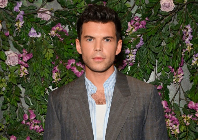 Luke Newton at an event, wearing a structured suit over a casual shirt, standing in front of a floral backdrop