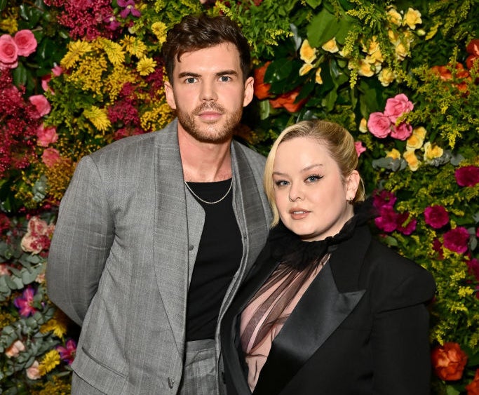 Nicola Coughlan in a black suit and sheer top, and Luke Newton in a gray suit with a black top, stand in front of a floral background