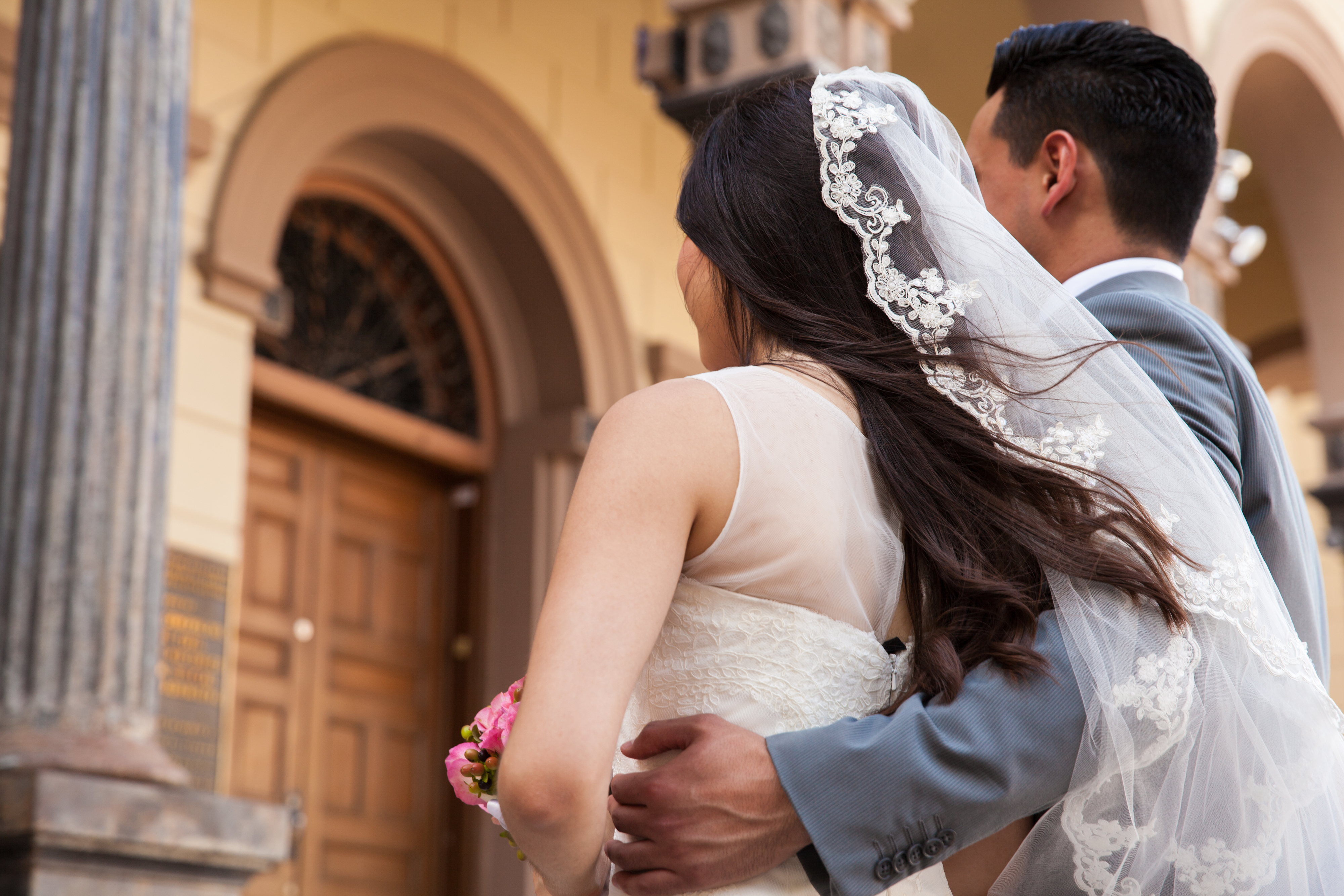 A bride in a lace veil and groom in a suit stand arm-in-arm outside a building with arched doorways