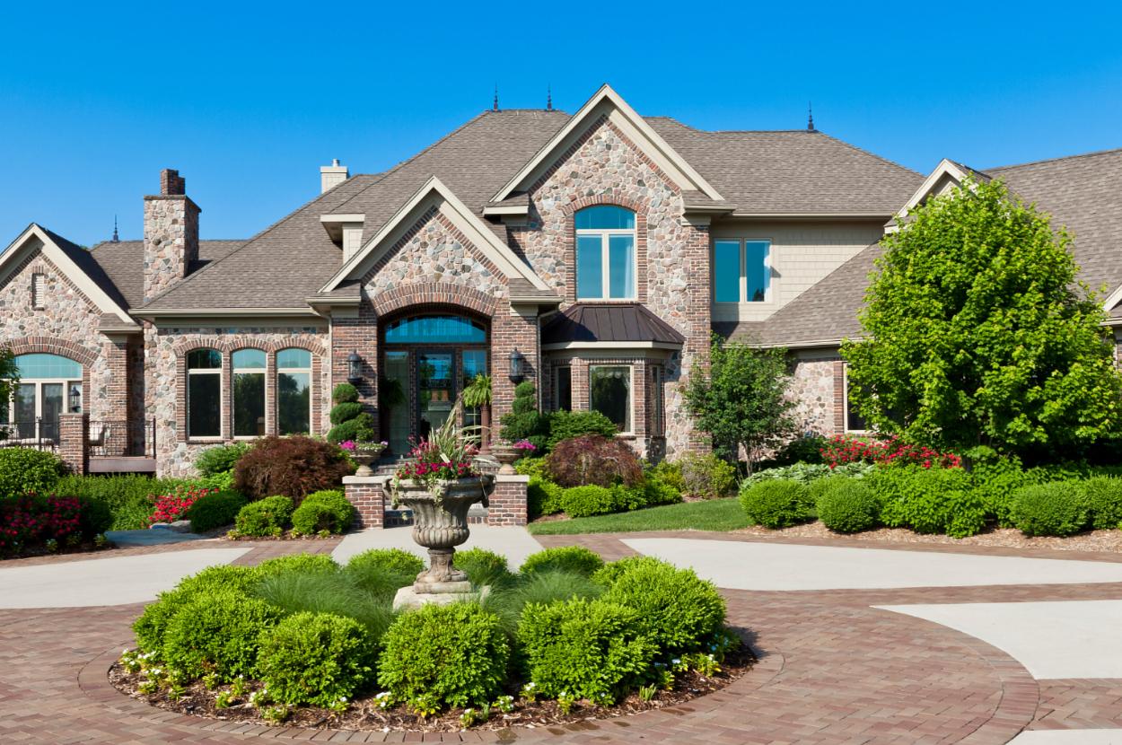 A large, elegant brick mansion with multiple gables and expansive windows, surrounded by well-manicured gardens and a central decorative fountain