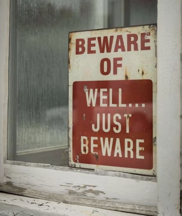 A sign in a window reads: "Beware of well… just beware." The sign is weathered and placed indoors
