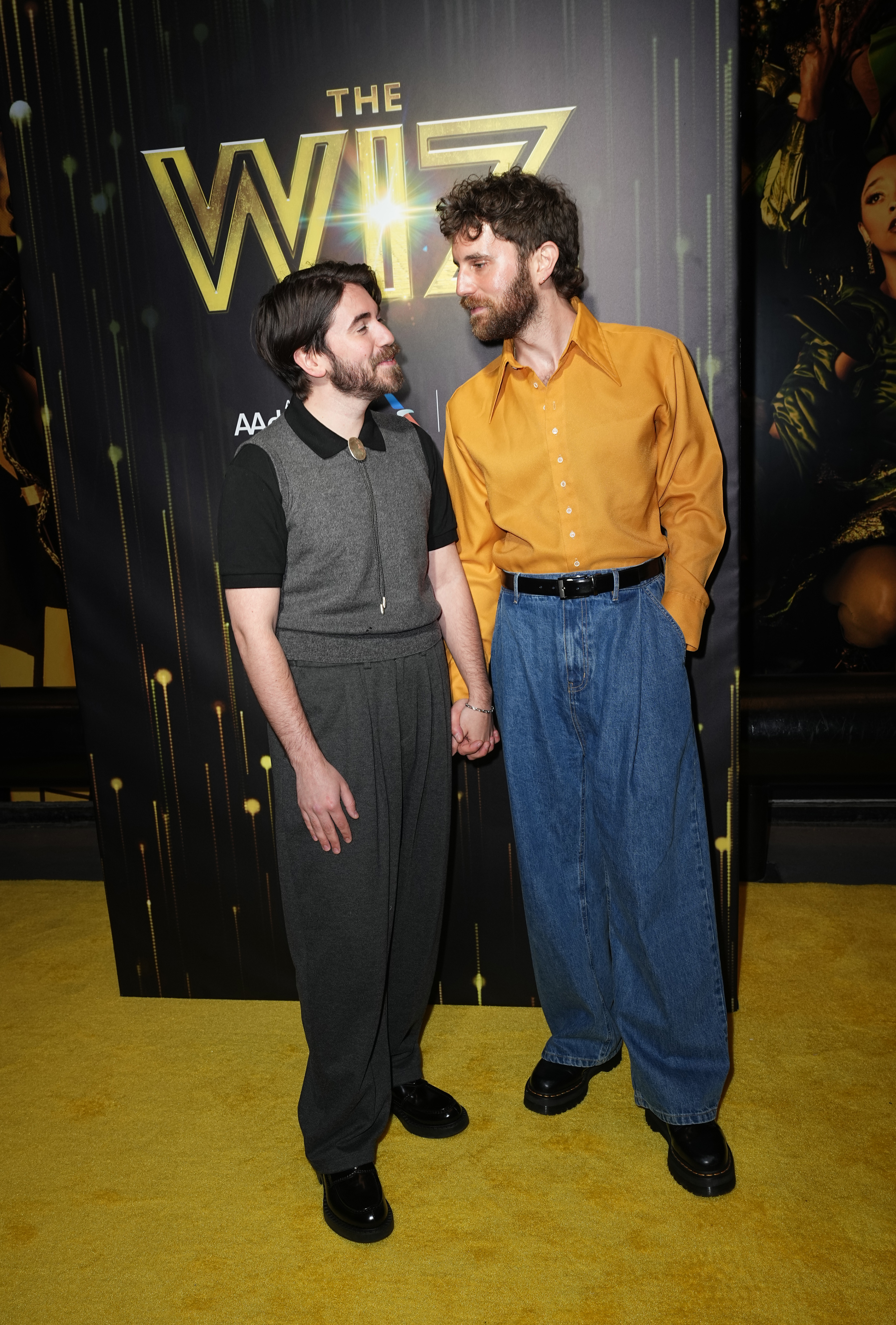 Two persons holding hands at "The Wiz" event. One in a gray and black outfit, the other in a yellow shirt and blue pants, standing on a yellow carpet