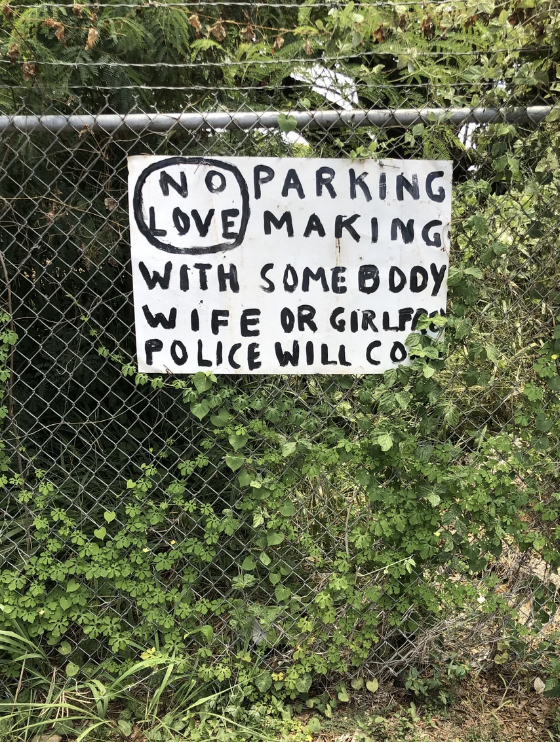 Sign on a chain-link fence reads "No parking lovemaking with somebody wife or girlfriend police will come."