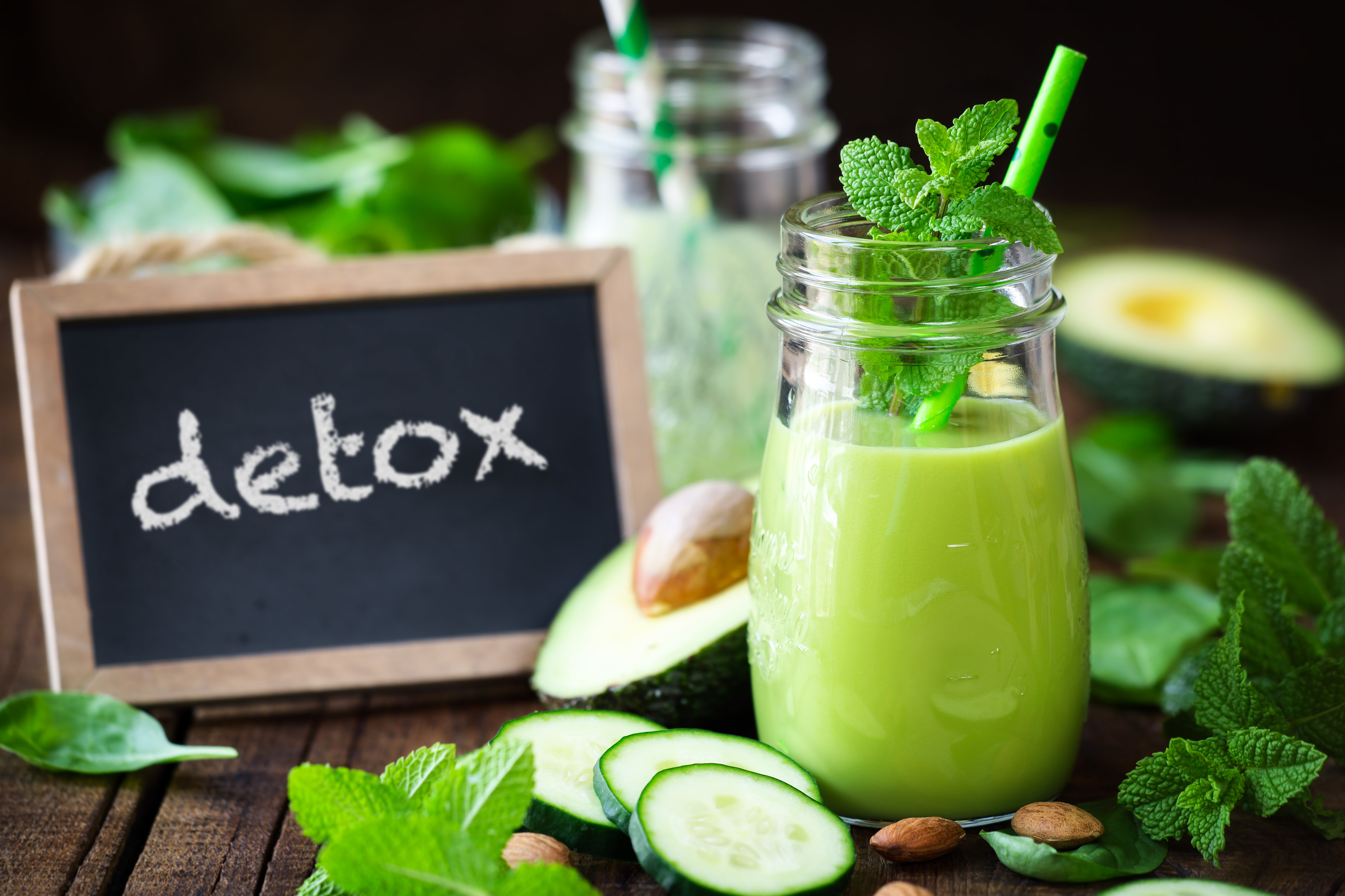 Two jars of green smoothie with mint leaves on top, surrounded by cucumber slices, avocado, spinach, and almonds; a small chalkboard with the word "detox" written on it