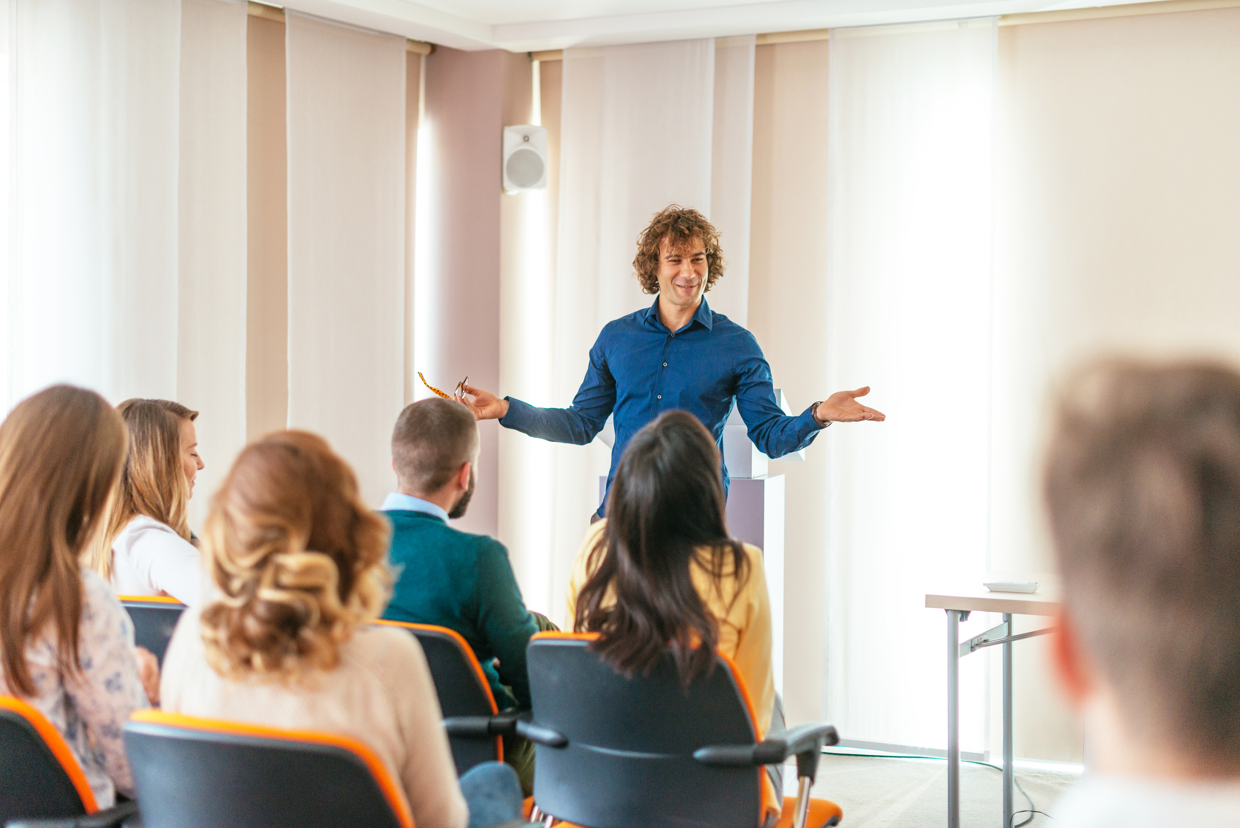 A man stands in front of a classroom, gesturing with his arms open, while several people sitting in chairs attentively listen