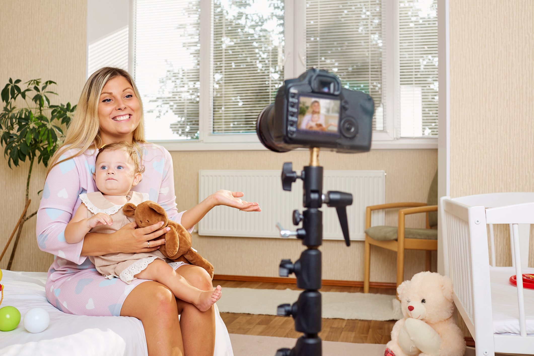 Adult sitting with a baby on a bed, looking at a camera on a tripod. They are surrounded by toys, including a teddy bear. The adult is smiling and gesturing