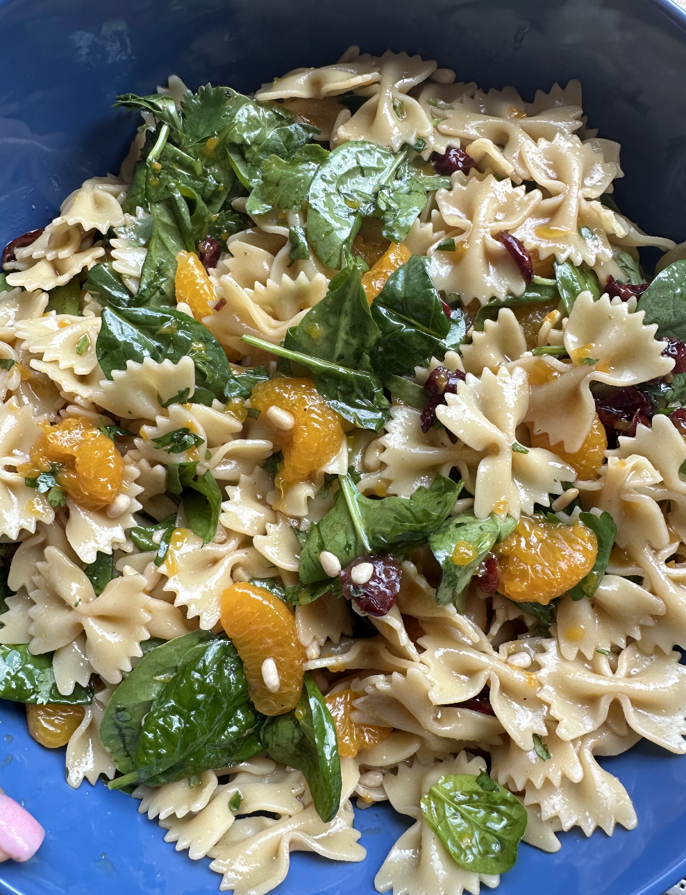 A bowl of pasta salad with farfalle pasta, spinach leaves, mandarin orange segments, dried cranberries, and pine nuts