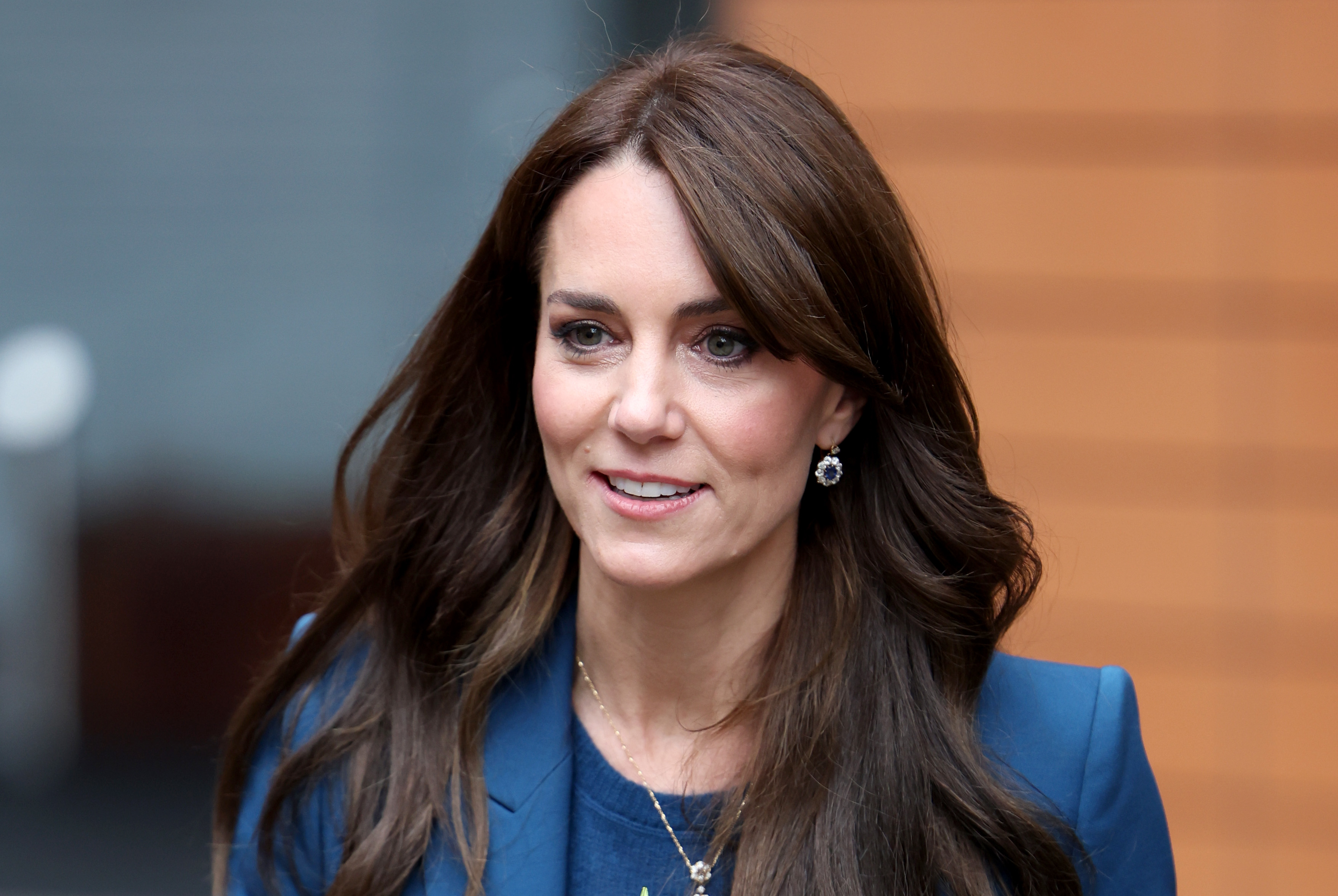 Kate Middleton smiles while attending a public event, wearing a blazer and matching top, with her hair styled down and simple earrings