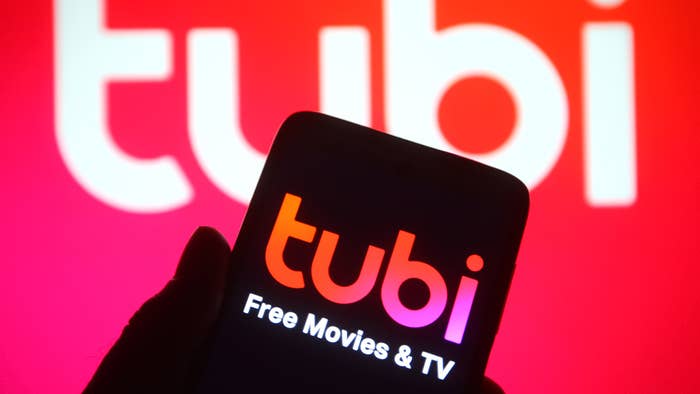 A hand holds a smartphone displaying the Tubi app, which has the text &quot;tubi Free Movies &amp; TV&quot; on the screen. In the background, the word &quot;tubi&quot; is prominently displayed