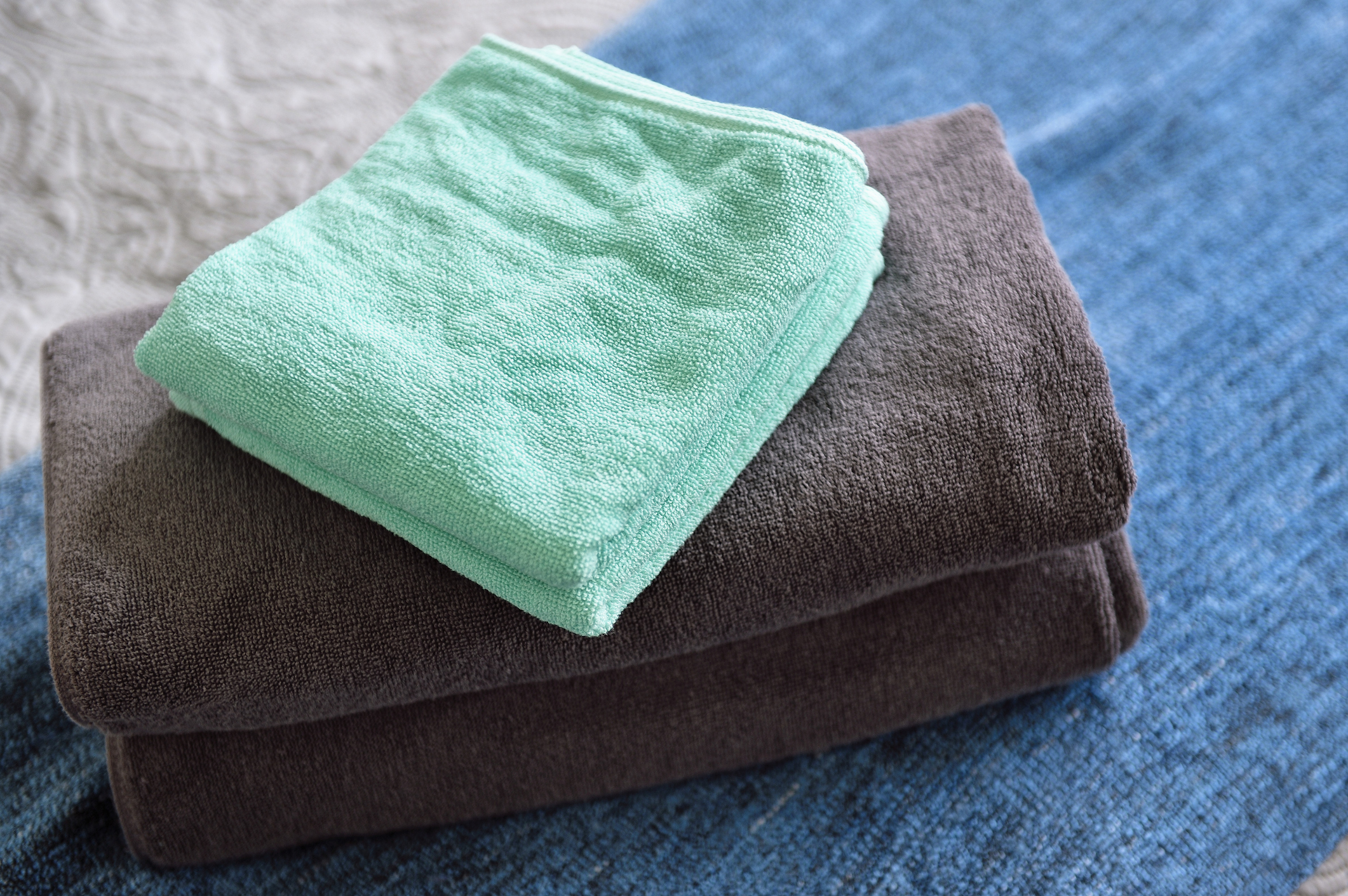 Stacked towels with a folded turquoise towel on top of two folded grey towels, placed on a blue surface