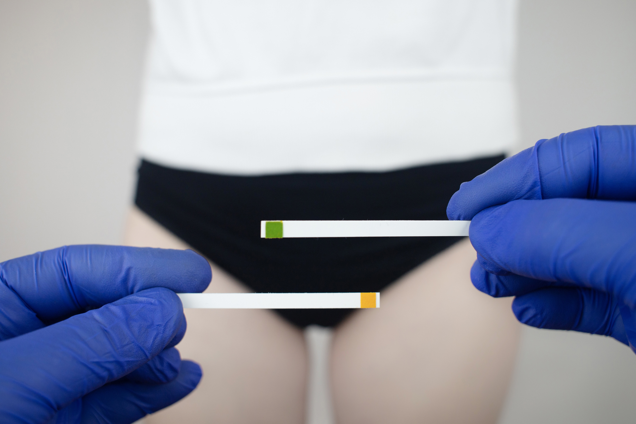 A person holding two pH testing strips, one green and one yellow, near their waist