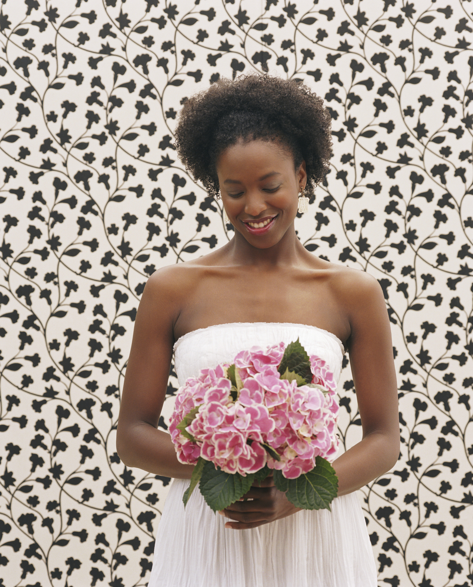 Woman smiles warmly, holding pink hydrangeas in front of a floral-patterned background