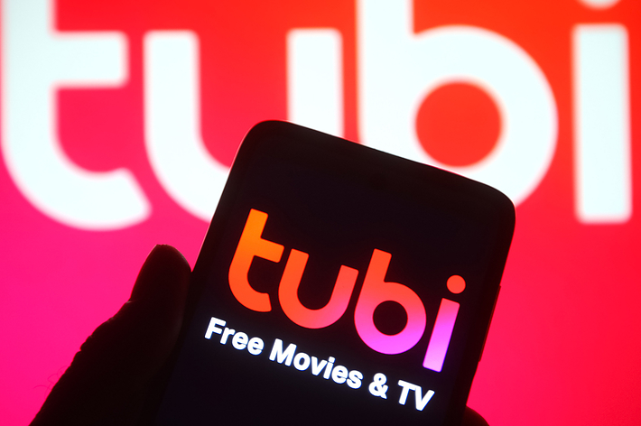 A hand holds a smartphone displaying the Tubi app, which has the text "tubi Free Movies & TV" on the screen. In the background, the word "tubi" is prominently displayed