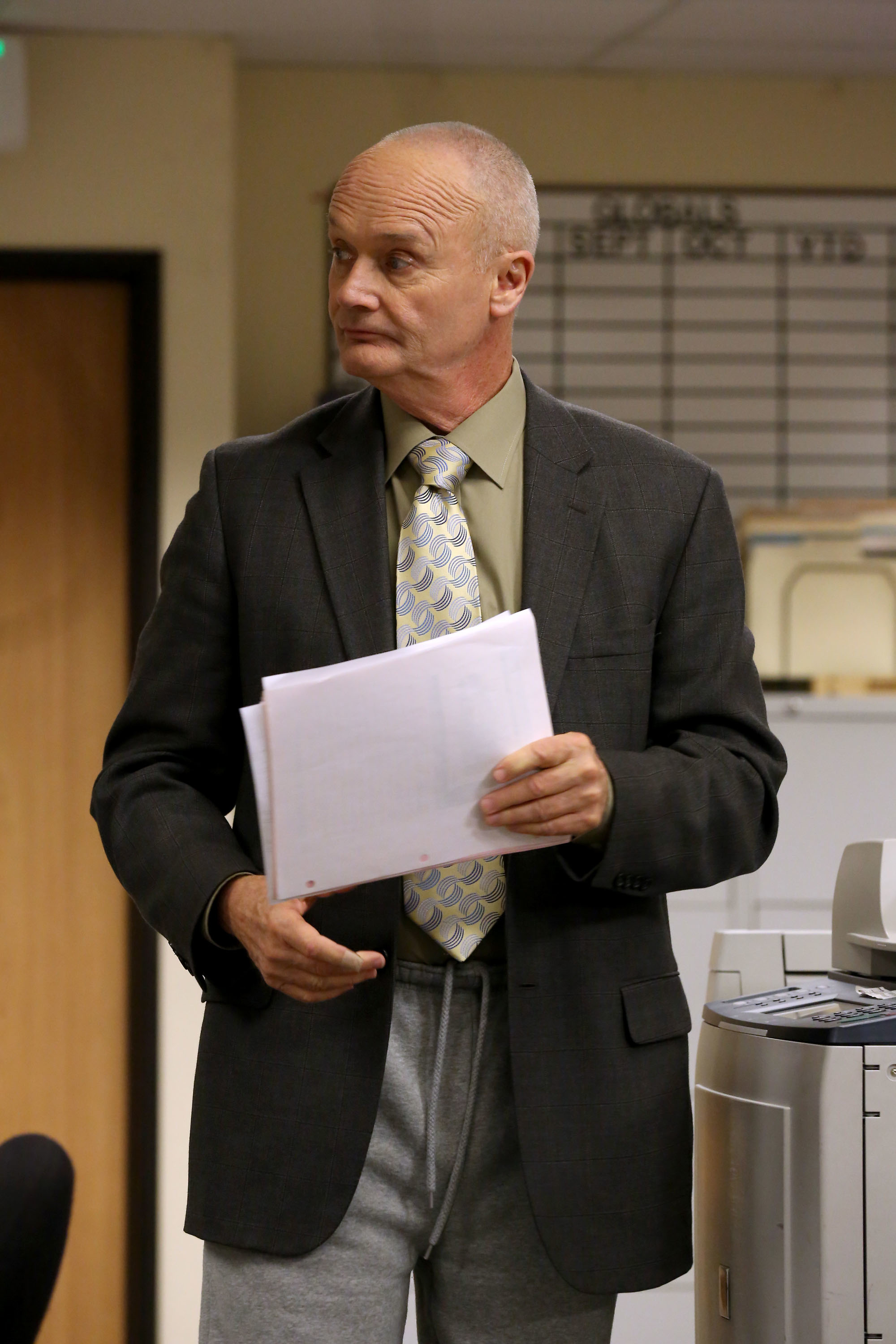 Creed Bratton wearing a suit jacket, tie, and sweatpants, holding papers, looking sideways in an office