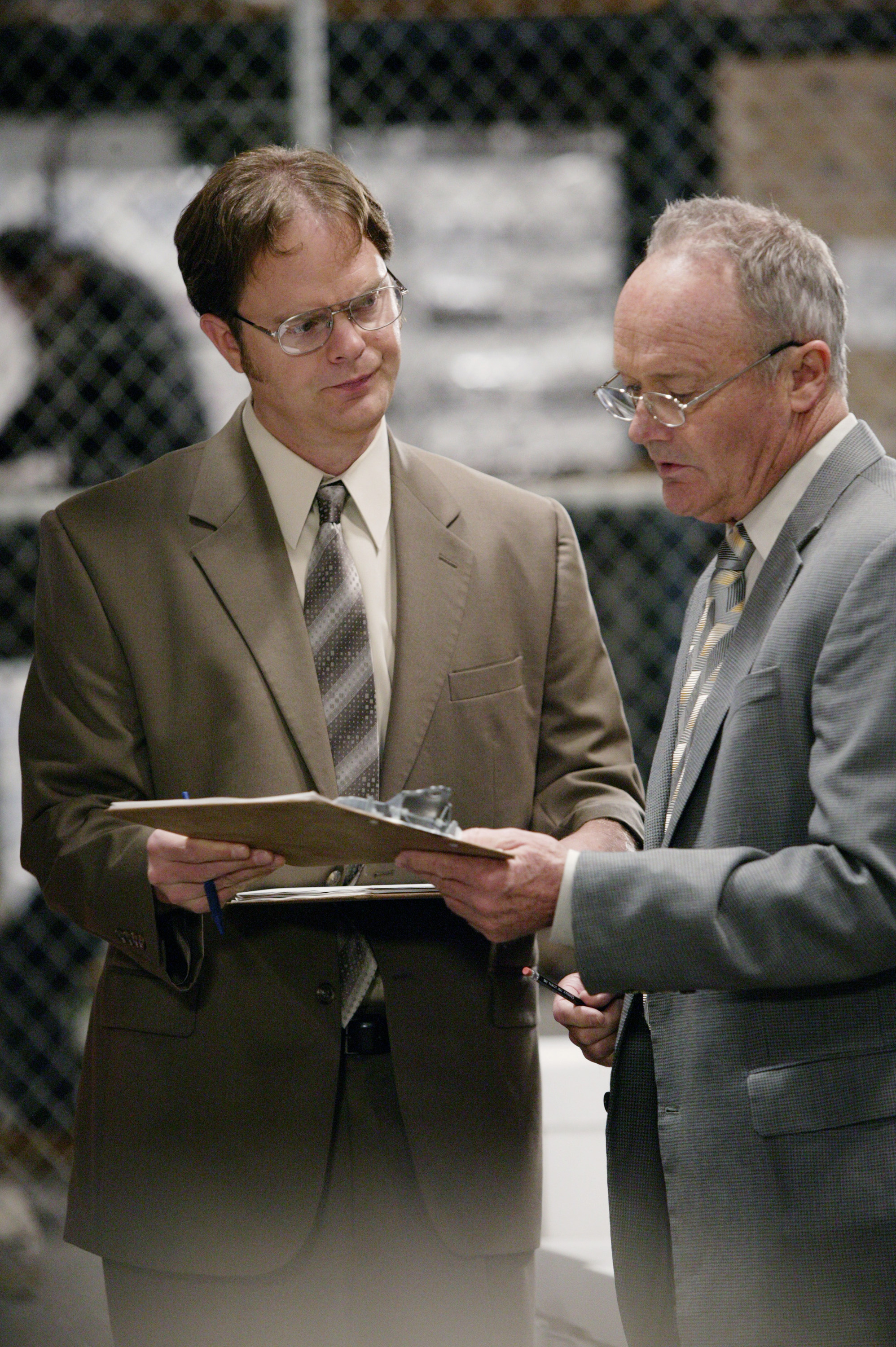 Rainn Wilson and Creed Bratton, both in suits, discuss documents on clipboards in an office