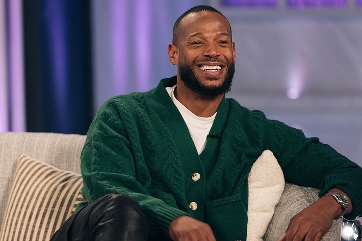 Marlon Wayans sits on a couch smiling, wearing a green cardigan and black pants.

