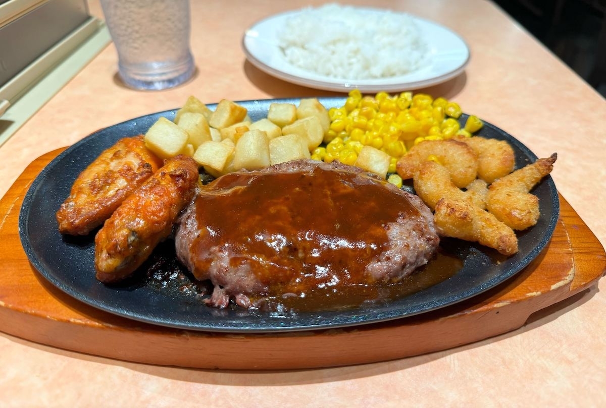 ステーキ、エビフライ、ポテト、コーン、唐揚げとご飯が並んでいる鉄板の食事がテーブルに置かれている。