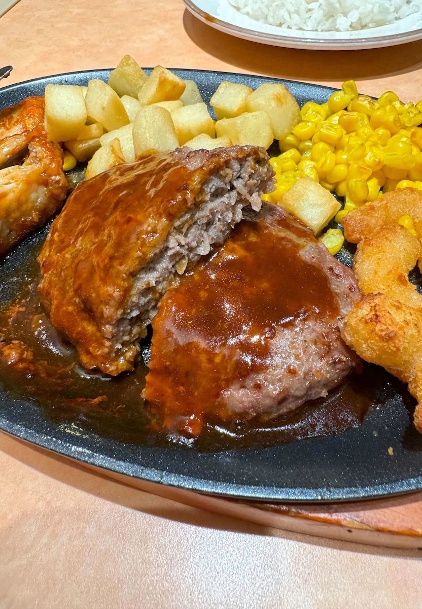 鉄板に盛られたハンバーグ、ポテト、コーン、エビフライの料理。後ろにはご飯の皿があります。