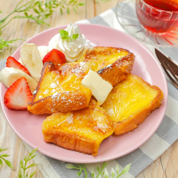 ピンクの皿に盛られたフレンチトースト、いちご、バナナ、ホイップクリームとバター付き。背景にナイフとフォーク。