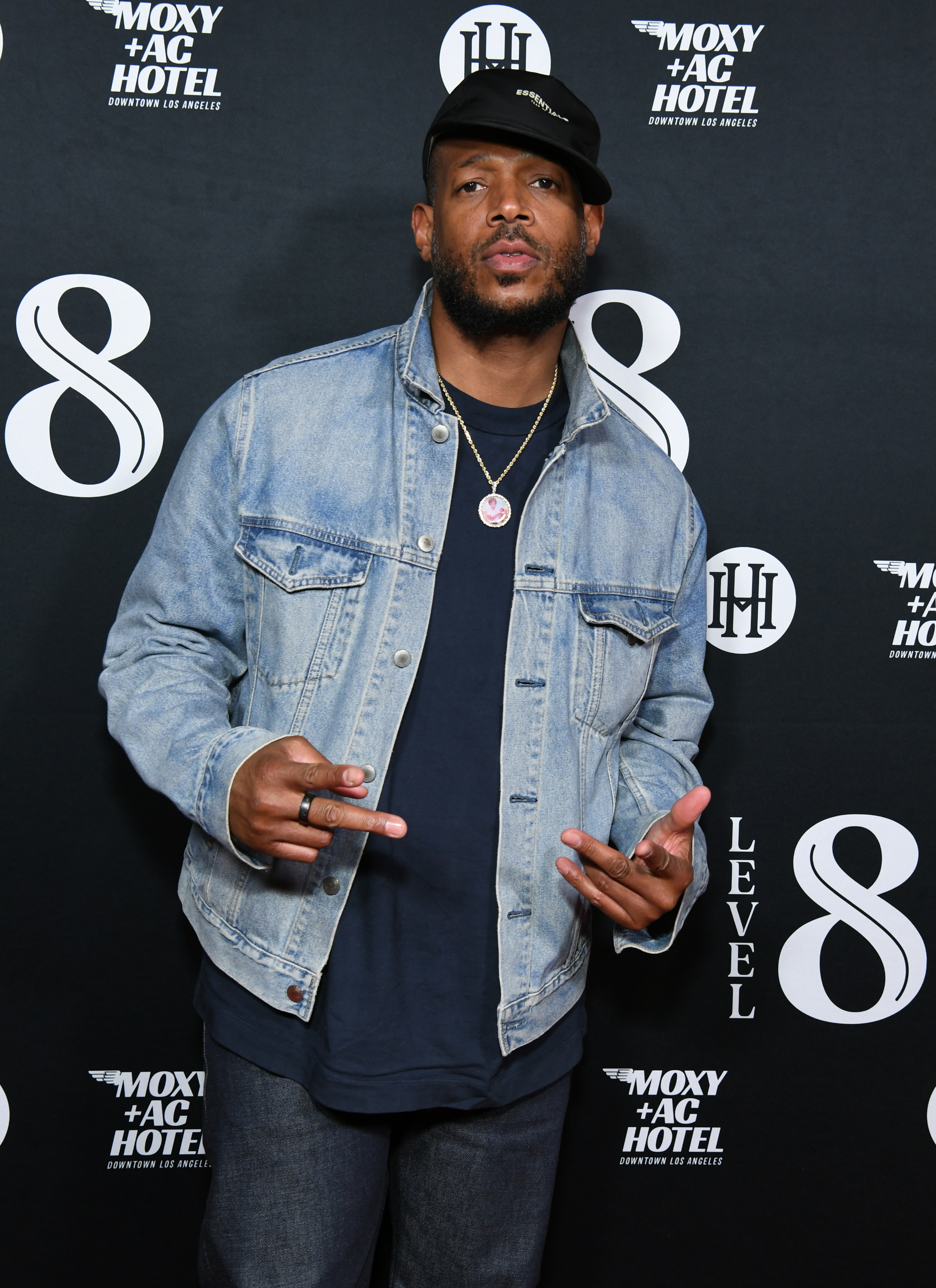 Marlon Wayans, in a denim jacket and black cap, poses with hand gestures at a Moxy + AC Hotel event in Los Angeles