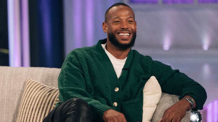 Marlon Wayans sits on a couch, wearing a green cardigan and black pants, smiling during an interview
