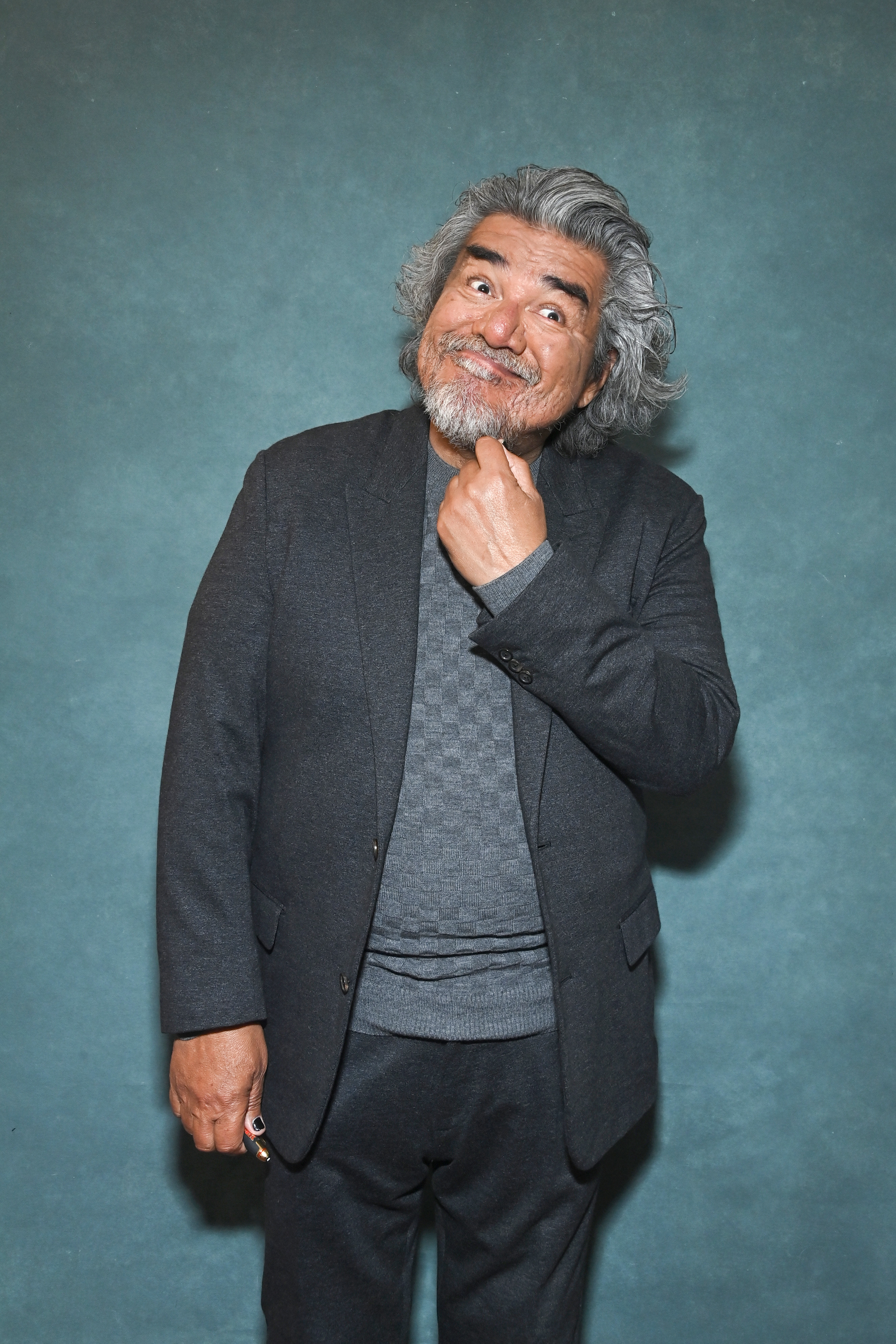 Jorge Perugorría stands against a plain background, wearing a dark suit and a sweater, with a thoughtful expression and his hand on his chin