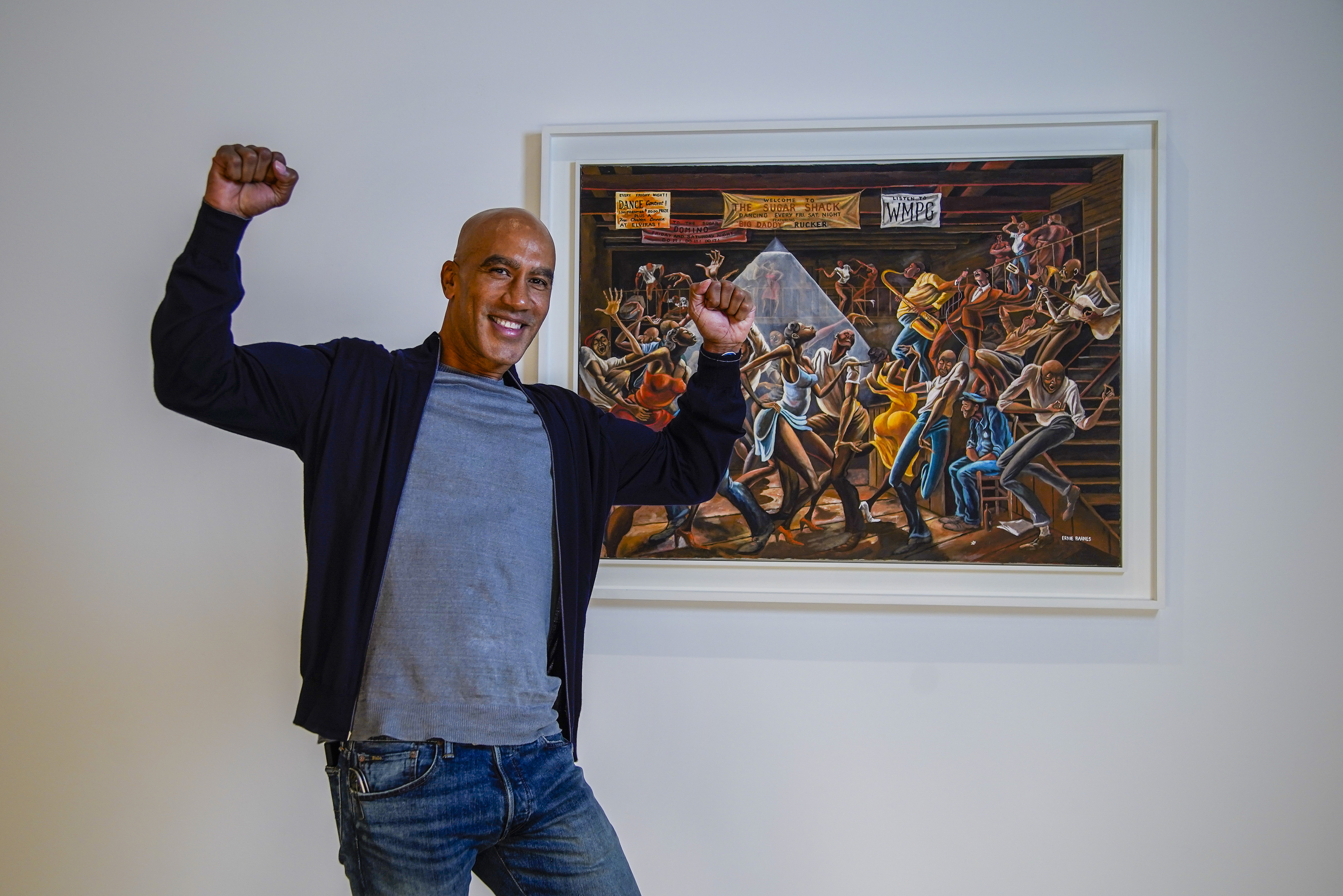 Man standing and smiling with arms raised in front of a painting depicting a lively dance scene