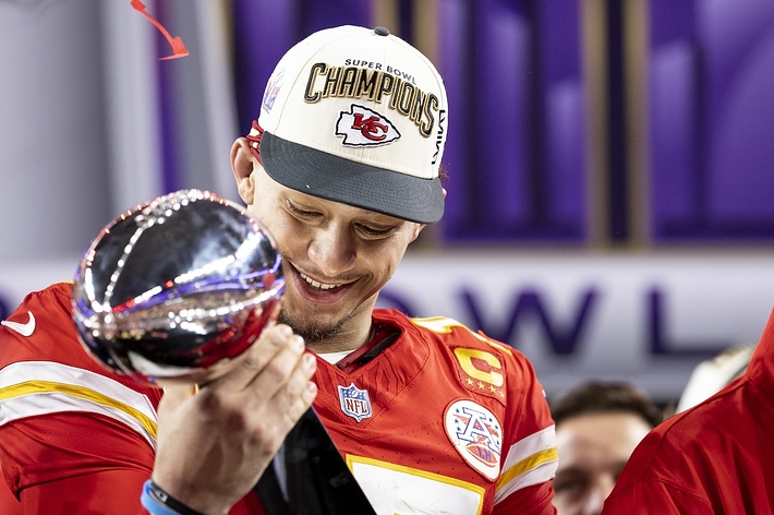 Patrick Mahomes in football uniform, wearing a "Super Bowl Champions" cap, smiles while holding the Vince Lombardi Trophy on a podium