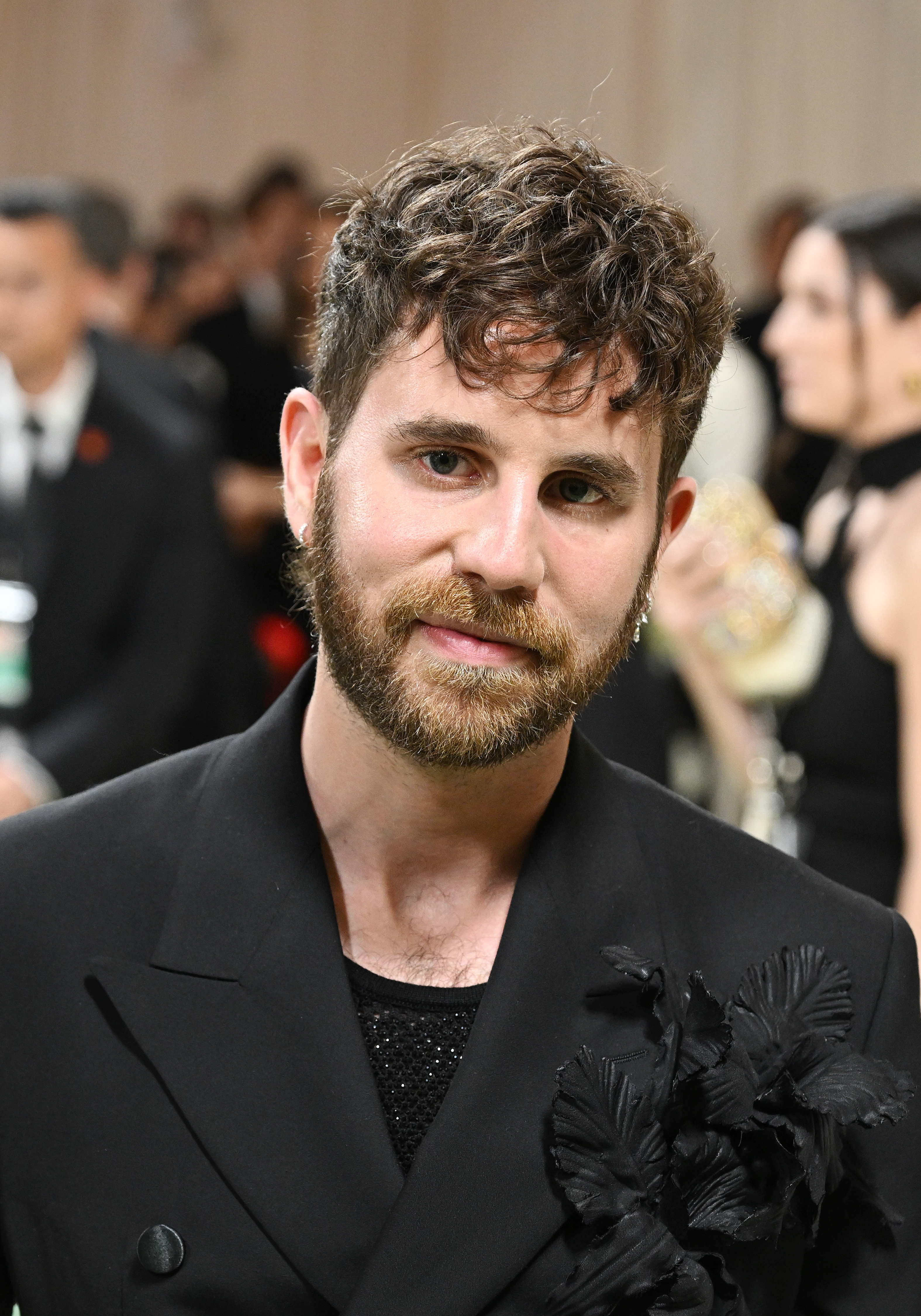Ben Platt in a stylish black suit with floral appliqué, smiles at the camera at an event with many attendees in the background