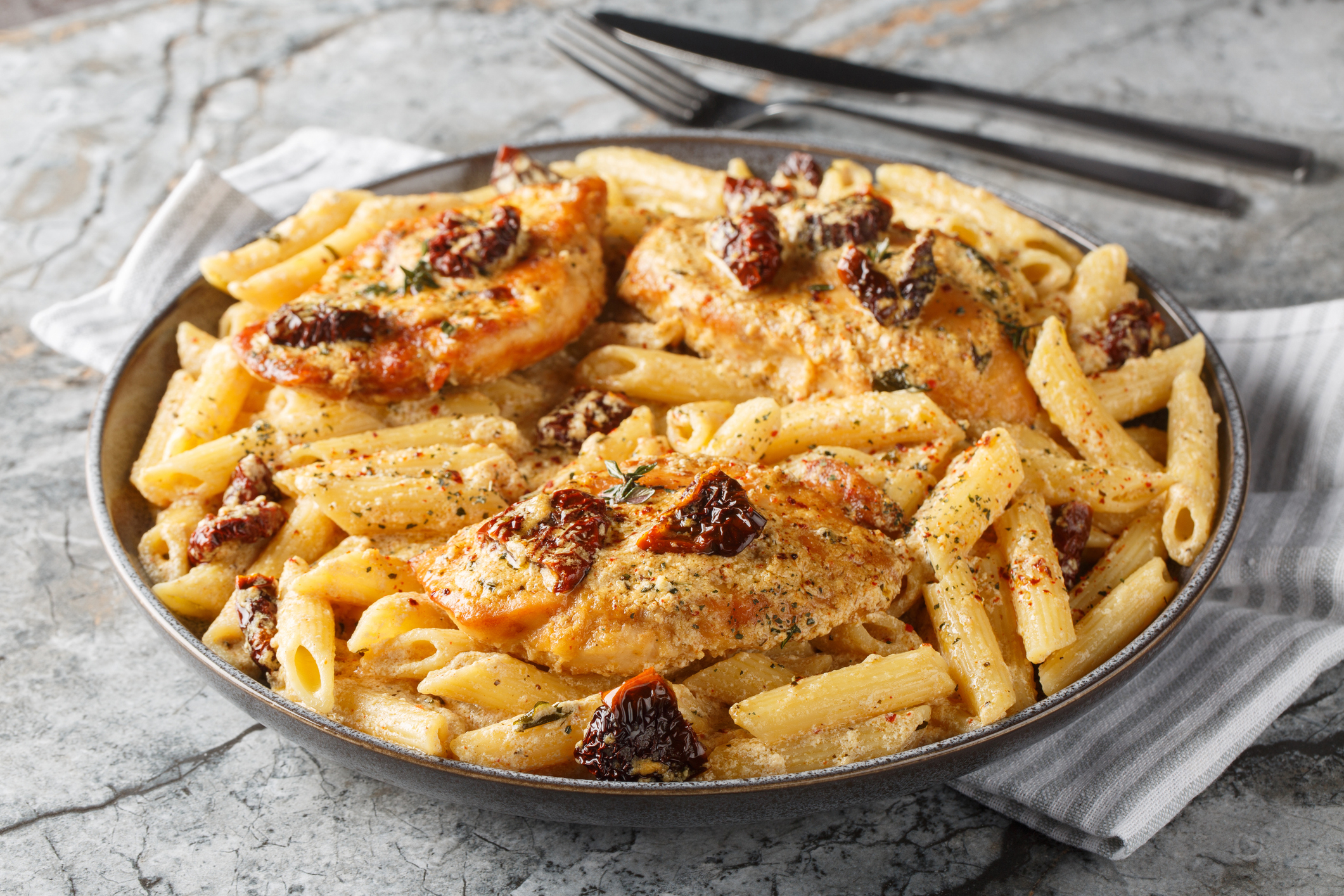A plate of creamy chicken pasta with penne, sun-dried tomatoes, and herbs on a textured surface, with a fork and knife on the side