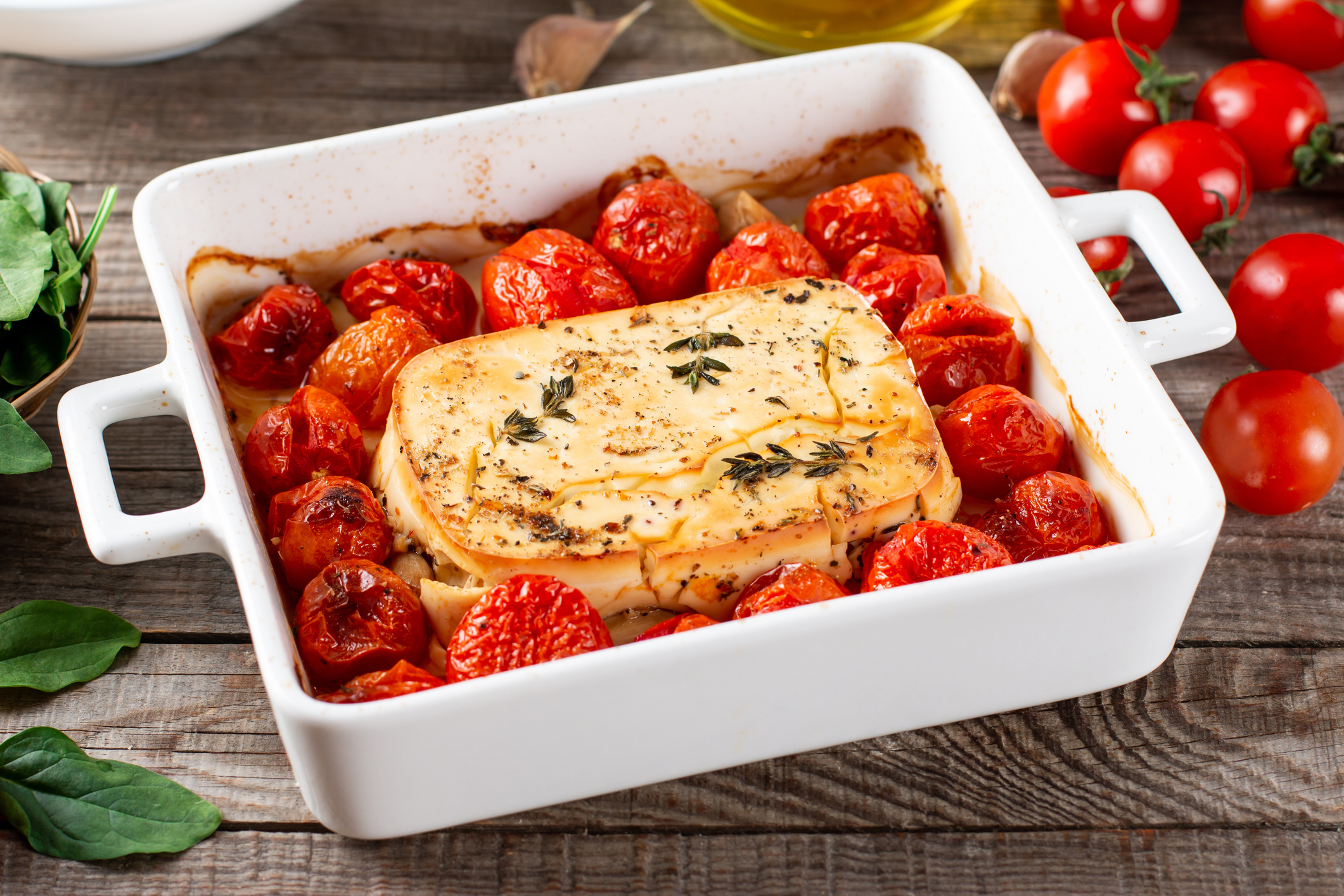 Baked feta cheese topped with herbs in a white dish surrounded by roasted cherry tomatoes on a wooden table