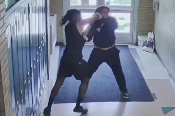 廊下で黒い服を着た2人の人物がもみ合っている監視カメラの映像。床には幾何学模様がある。