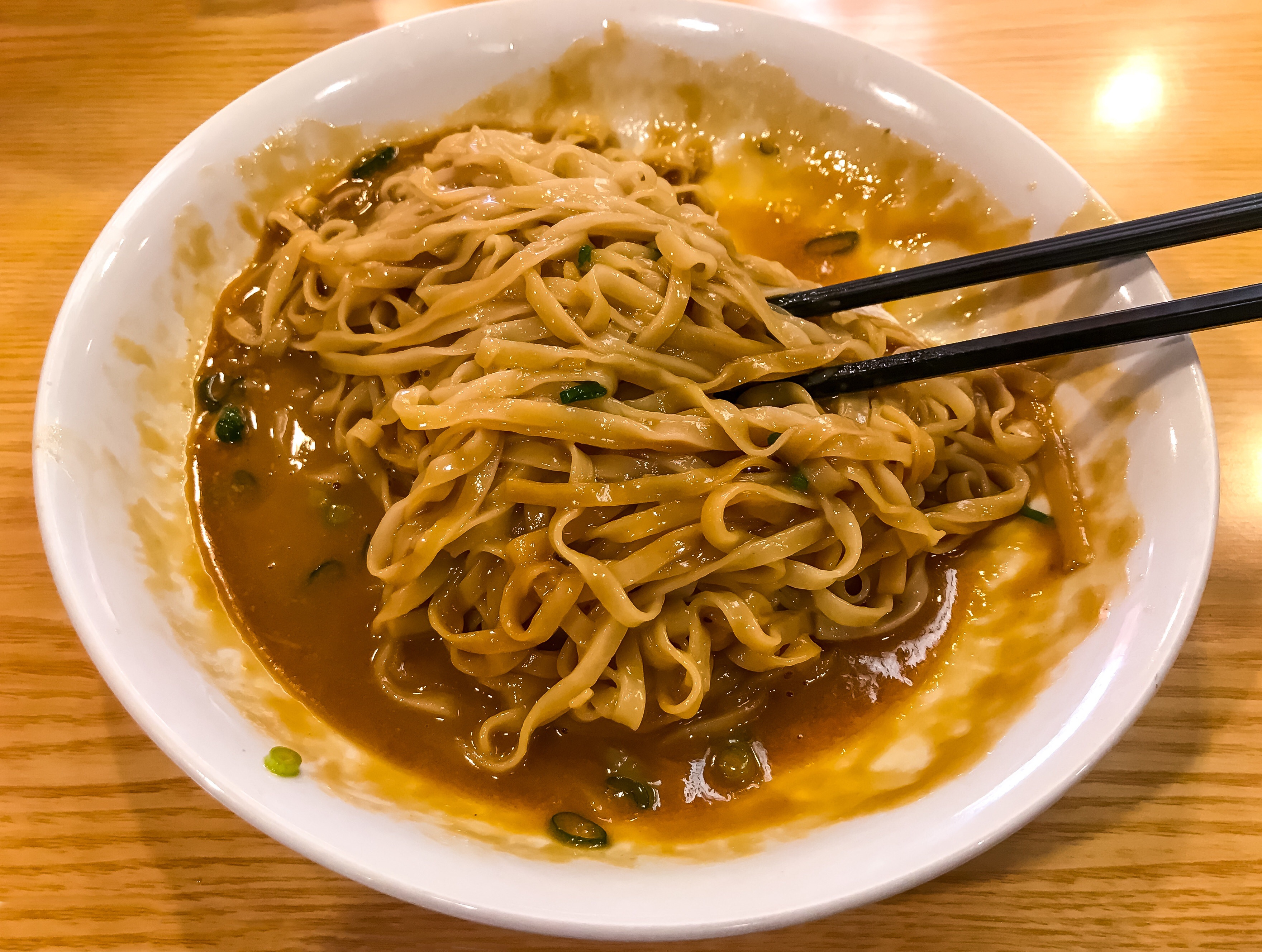 A bowl of noodles mixed in a sauce with green onions, partially eaten, and a pair of chopsticks resting in the bowl