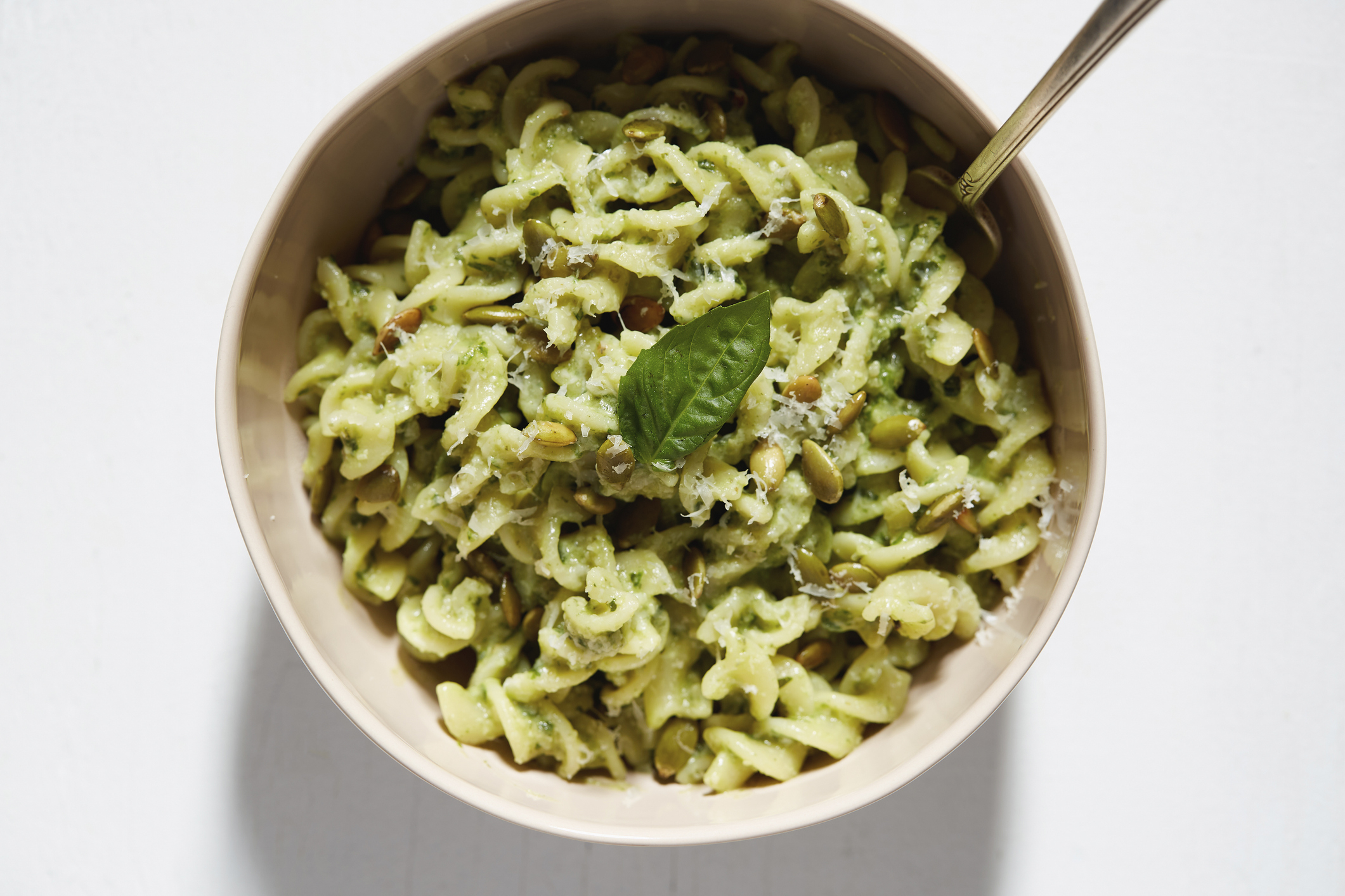 A bowl of pasta coated in a creamy green pesto sauce, garnished with a basil leaf and pine nuts. A spoon is placed in the bowl