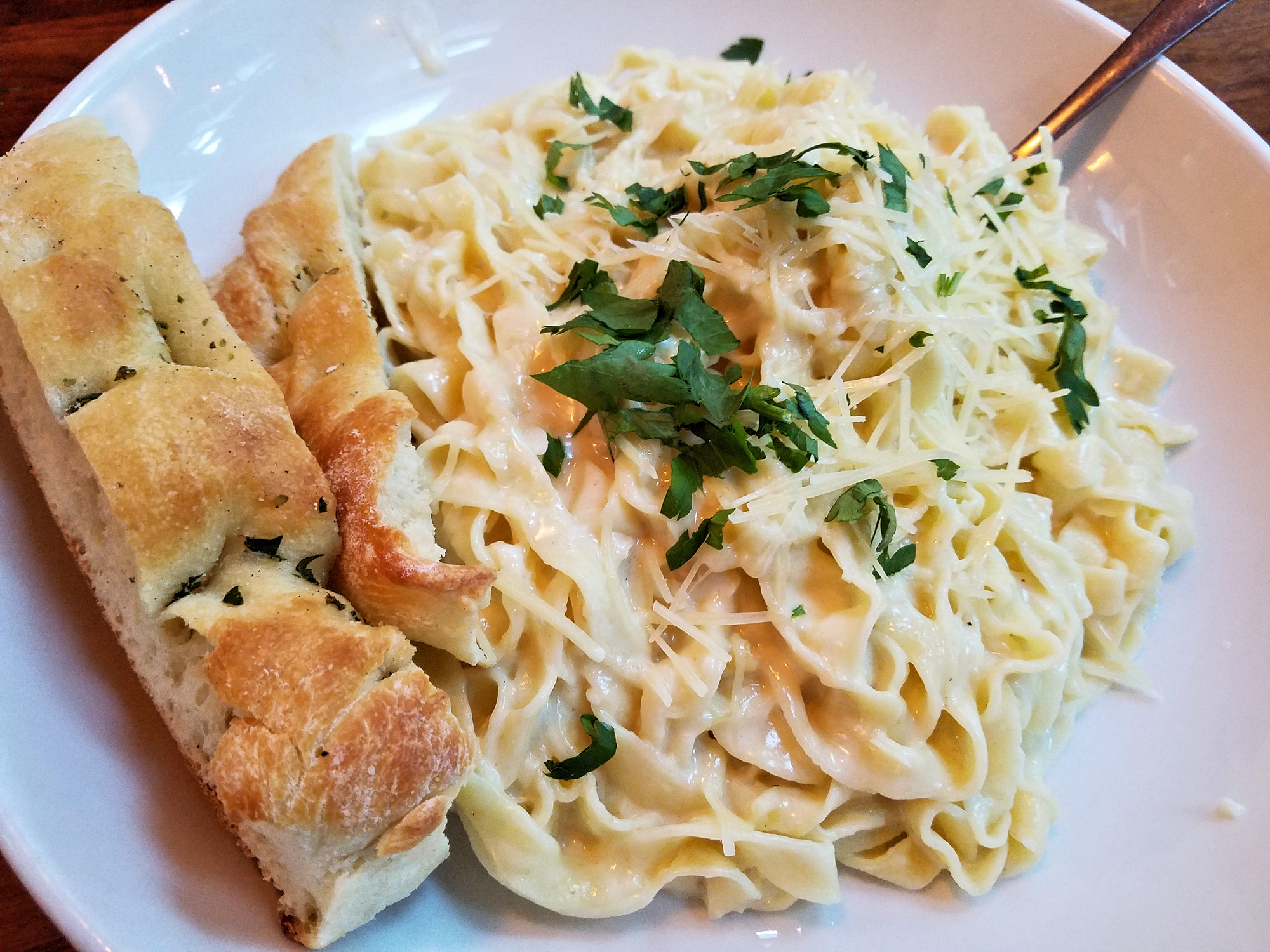 A plate of fettuccine Alfredo garnished with grated cheese and parsley, served with two slices of garlic bread and a fork