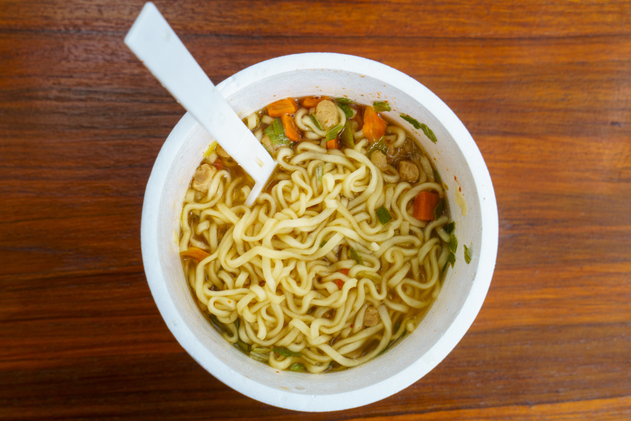 A bowl of instant noodles with vegetables on a wooden surface, with a white plastic spoon inserted into it
