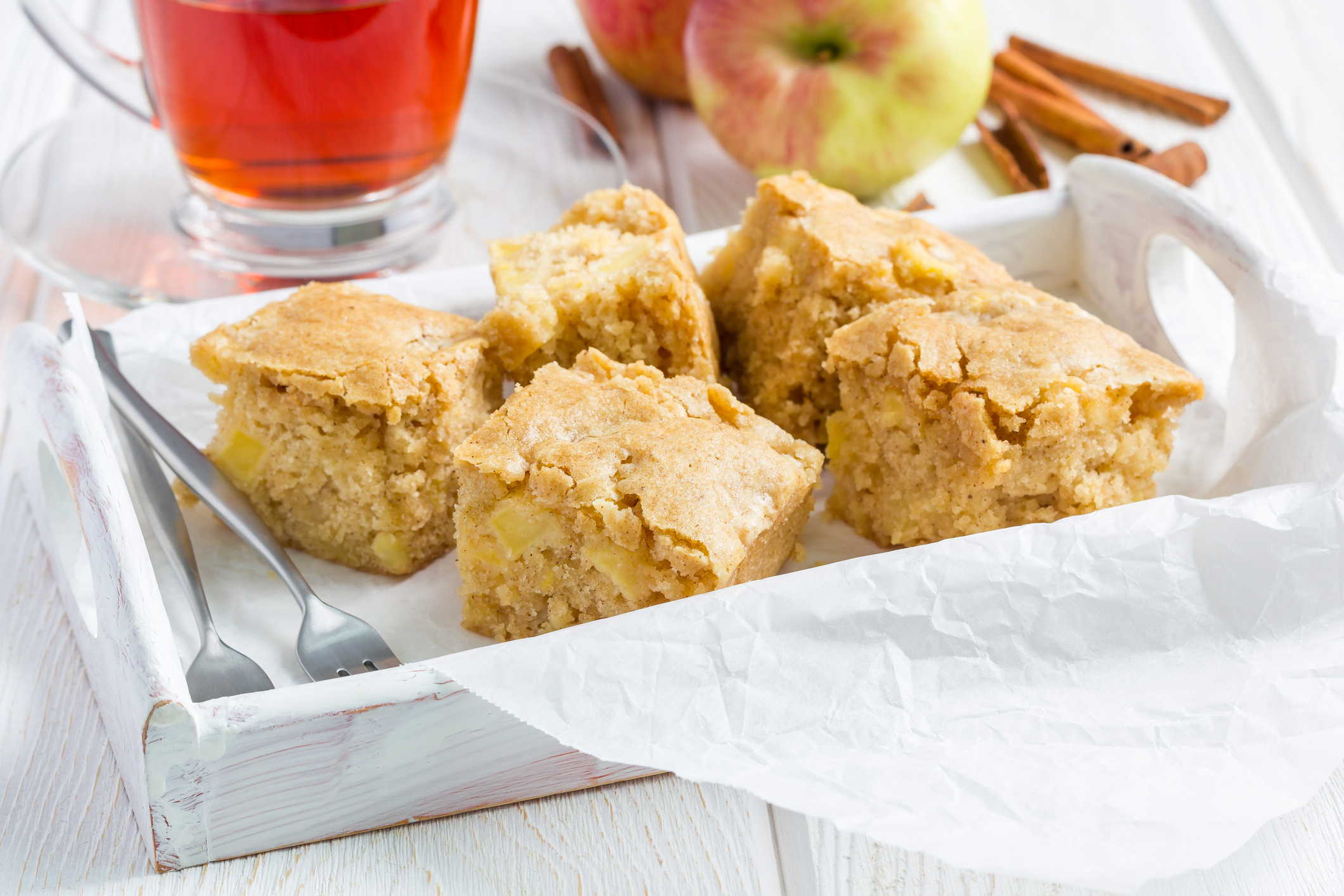 Tray with five apple cake squares, a glass of tea, two apples, and cinnamon sticks on a white wooden table