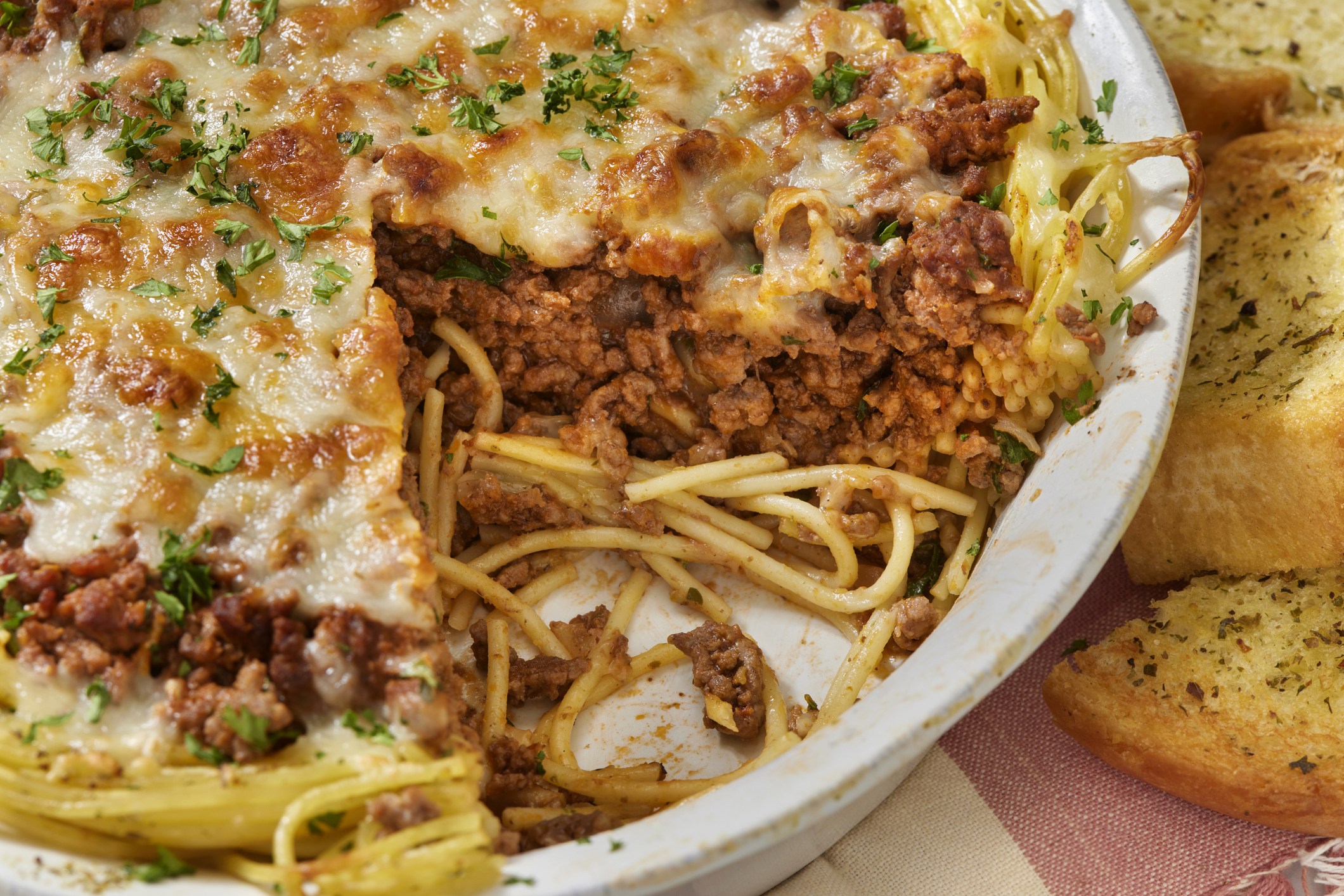 A dish of spaghetti pie with melted cheese and meat sauce, partially served, accompanied by slices of garlic bread