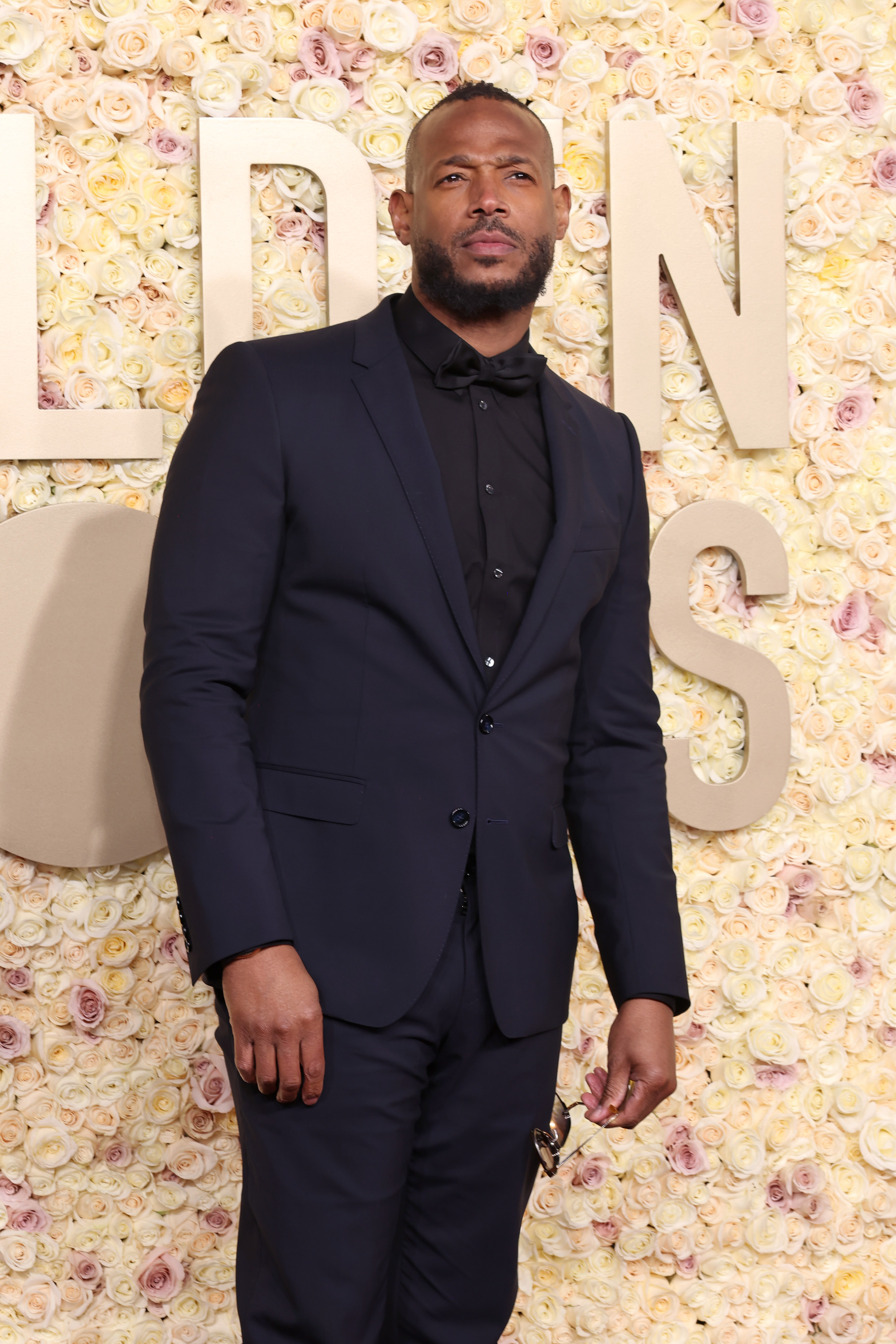 Marlon Wayans on the red carpet, wearing a black suit with a black dress shirt and bow tie. Background features floral arrangement and large text