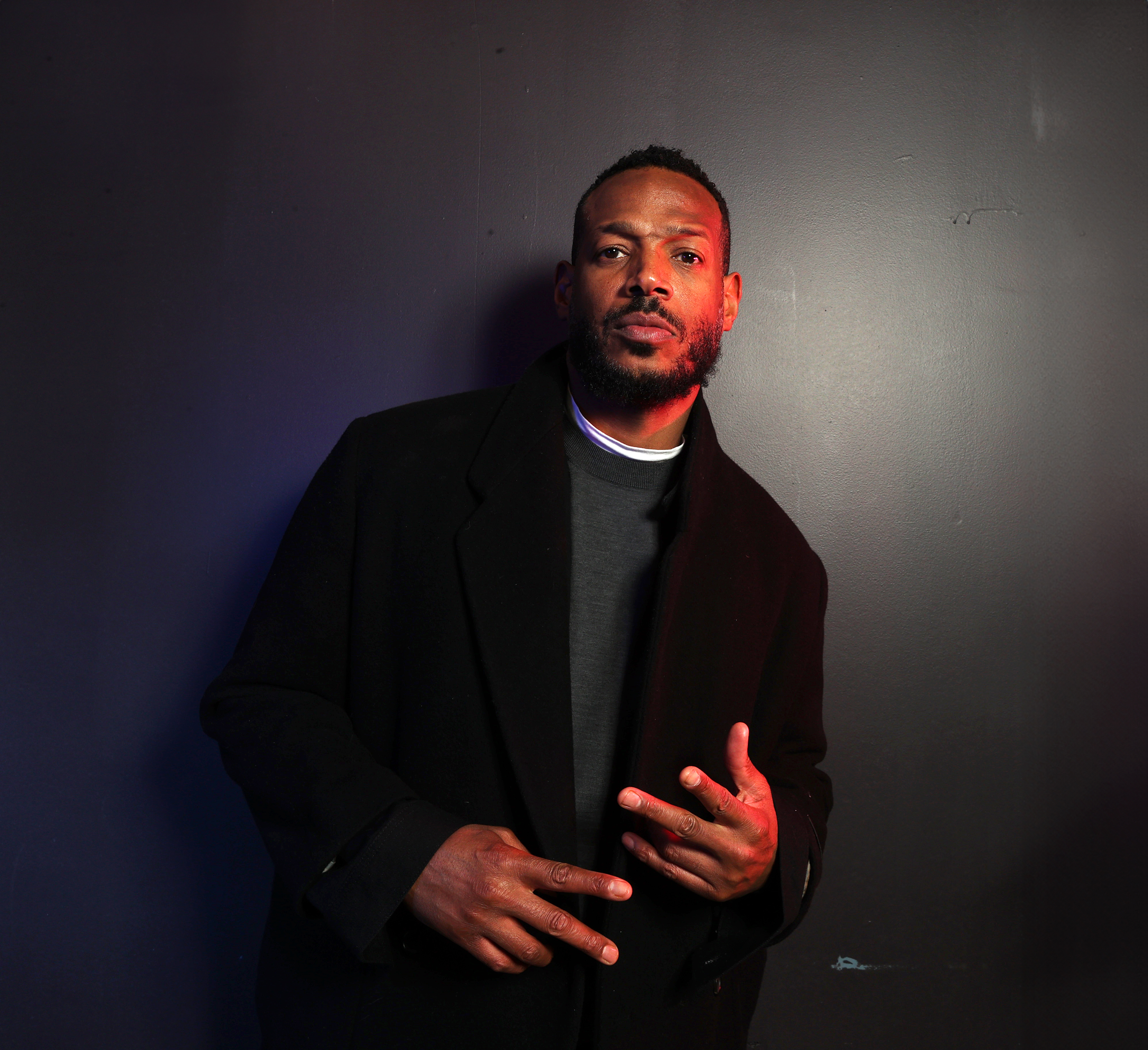 Marlon Wayans poses in a dark suit coat with a neutral expression, flashing a peace sign with his right hand