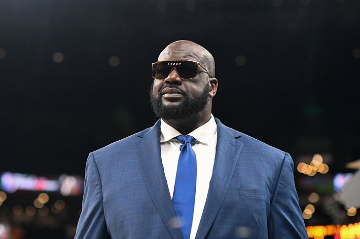 Shaquille O'Neal in a blue suit and tie, wearing sunglasses, standing in an indoor venue with blurred background lights