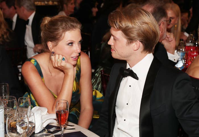 Taylor Swift and Joe Alwyn sit at a formal event, engaged in conversation. Taylor wears a stylish evening gown, while Joe is in a classic black tuxedo