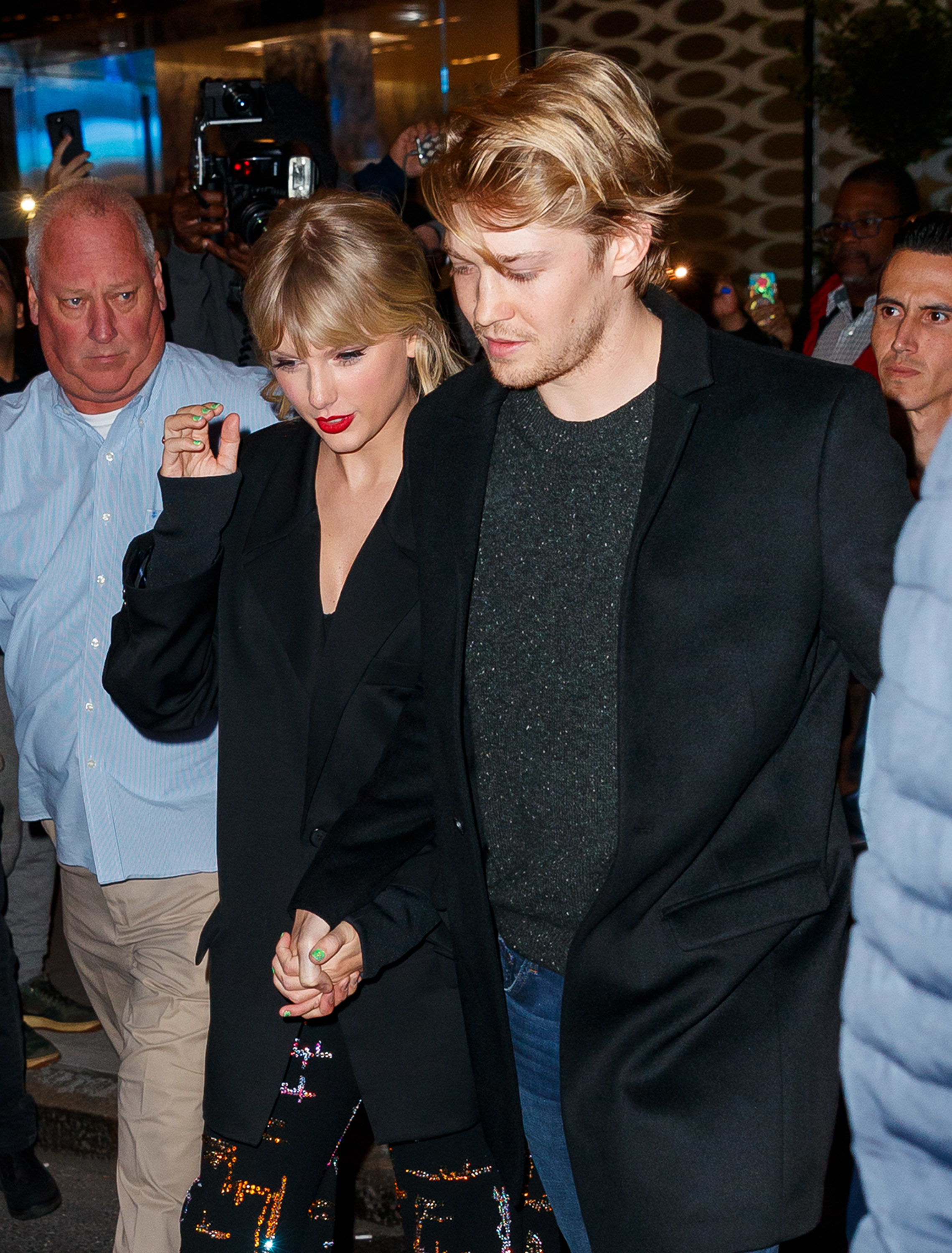Taylor Swift and Joe Alwyn hold hands while walking outside, surrounded by people. Taylor wears a black outfit with colorful patterns, and Joe wears a black jacket