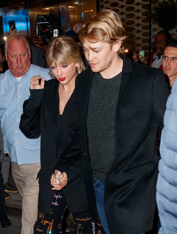 Taylor Swift and Joe Alwyn hold hands while walking outside, surrounded by people. Taylor wears a black outfit with colorful patterns, and Joe wears a black jacket