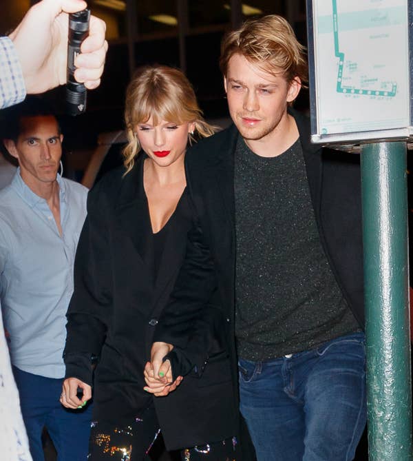 Taylor Swift, in a stylish outfit with a black blazer, holds hands with Joe Alwyn, wearing a dark sweater and jeans. They walk with another man in a light shirt