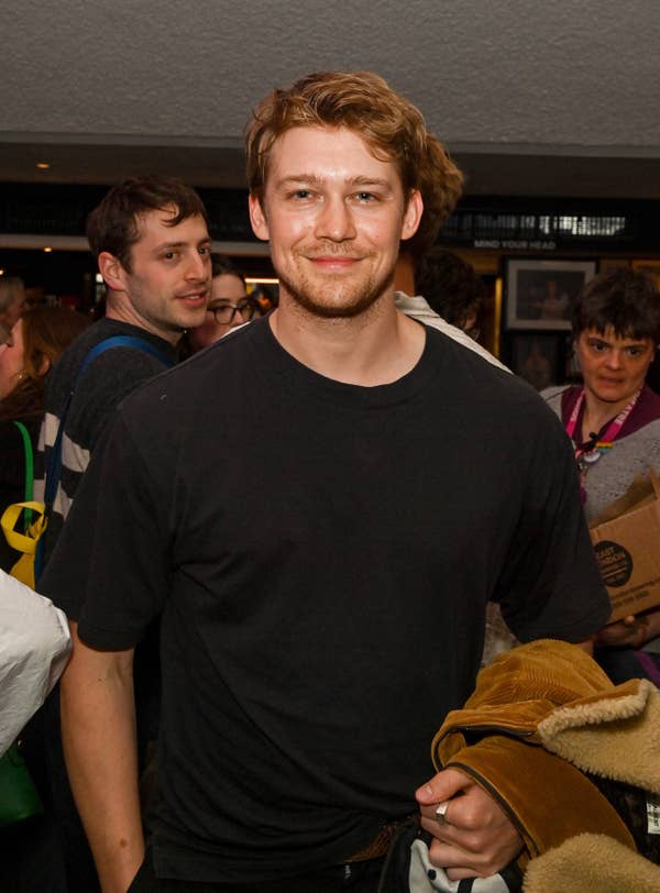 Joe Alwyn, wearing a casual black T-shirt, smiles at an event with people in the background